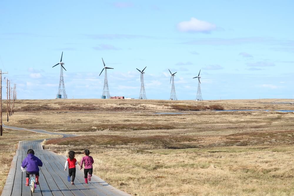Three children running towards wind turbines in the distance